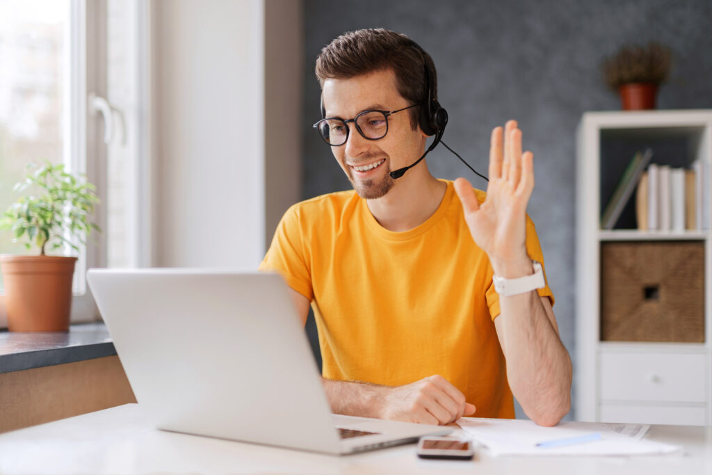 Man in smart orange sweatshirt waving to colleagues whilst working from home.
