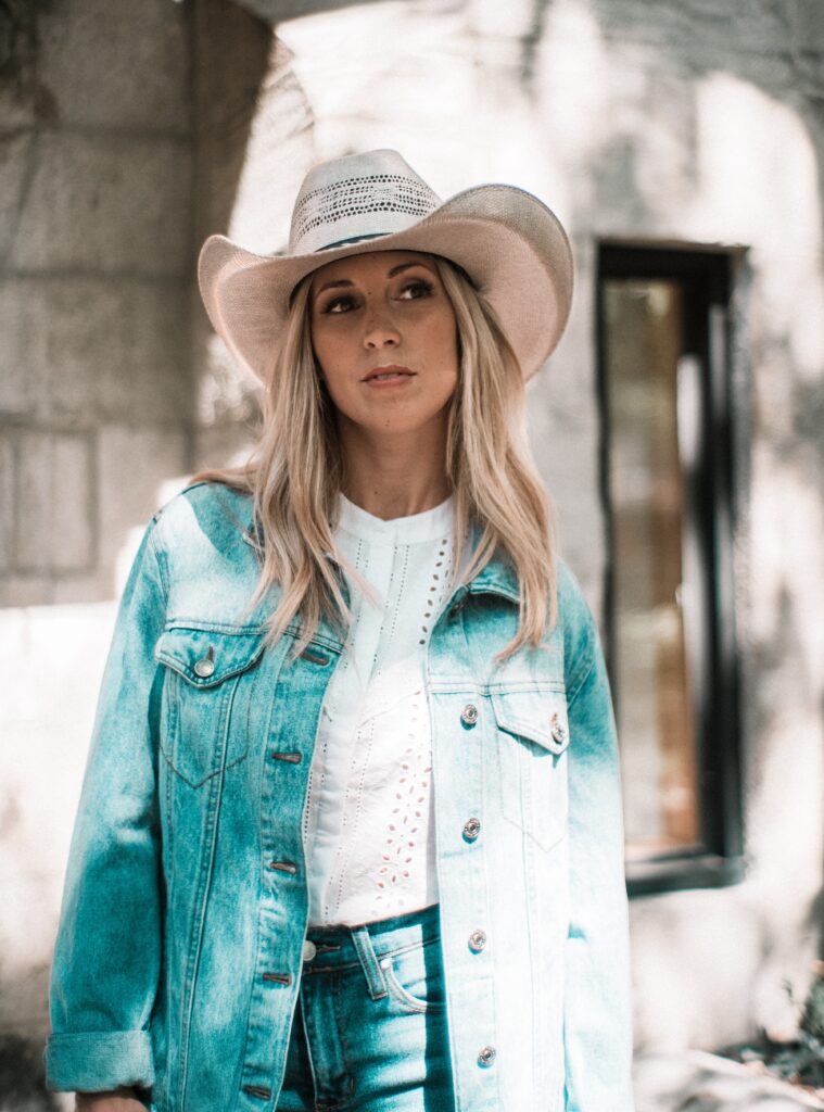 A lady wearing a cowgirl inspired outfit with a cowgirl hat, a coastal-style white dress, denim jeans and a denim jacket.